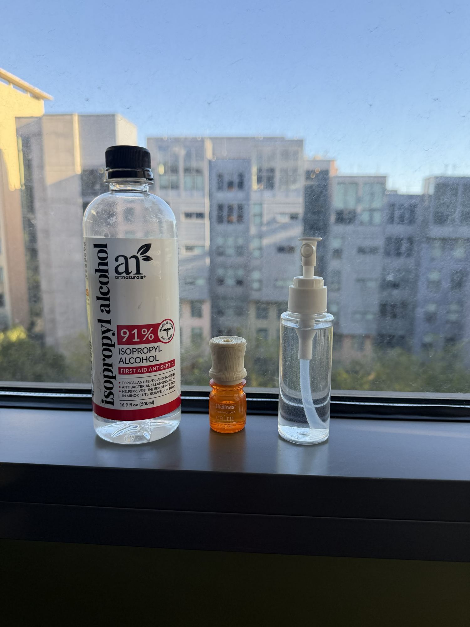 A bottle of 91% isopropyl alcohol, a small amber bottle of scented oil, and a clear spray bottle on a windowsill.